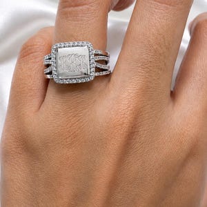 May include: A silver-toned ring featuring a square-shaped center plate engraved with the letters KKK. The plate is surrounded by a halo of small clear stones, and the band consists of three split rows also encrusted with small clear stones.