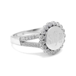 May include: A silver ring with a round, engraved center featuring the initials "SCK". The center is surrounded by a circle of small, clear stones.