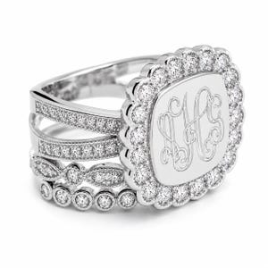 May include: A silver-toned ring featuring a central rectangular plaque engraved with the monogram AHS. The plaque is surrounded by a scalloped halo of small round stones. The band consists of three distinct rows encrusted with small stones.