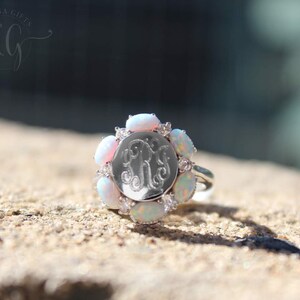 May include: A silver ring with a round, engraved monogram surrounded by a halo of white opals and small clear stones.