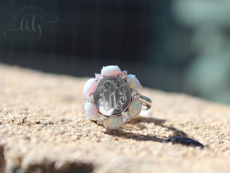 May include: A silver ring with a round, engraved monogram surrounded by a halo of white opals and small clear stones.