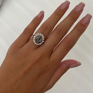 Könnte beinhalten: Ein silberfarbener Ring mit einer kreisförmigen Platte, in deren Mitte die Buchstaben FLB eingraviert sind. Die mittlere Scheibe ist von einem Kranz aus kleinen, klaren, rund geschliffenen Steinen in einer Metallfassung umgeben.