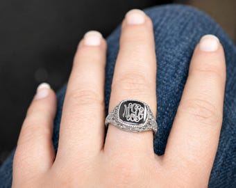 May include: A silver-toned ring featuring a square-shaped black inlay with an intricate, engraved white monogram. The band and setting display detailed, filigree-style metalwork with a textured, ornate pattern surrounding the central stone.