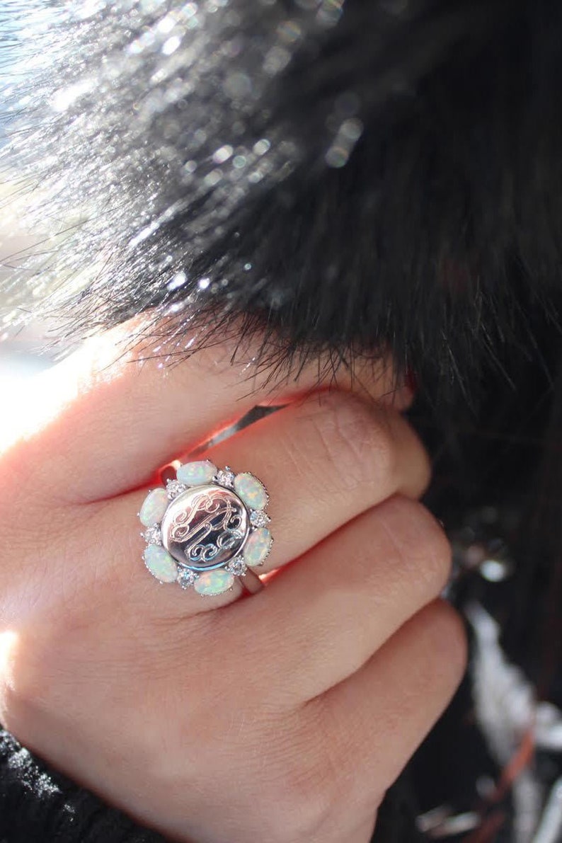 May include: A silver ring with a round opal stone and a monogram in the center. The ring is surrounded by smaller opal stones.
