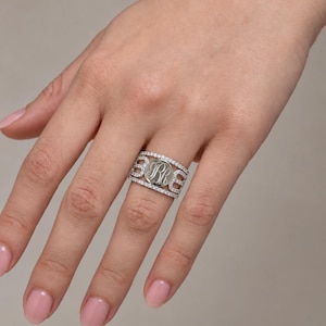 May include: A wide silver-toned ring featuring a central circular disc engraved with the letters RR. The band is composed of three horizontal rows of small, clear, sparkling stones, with the central disc integrated into the design.