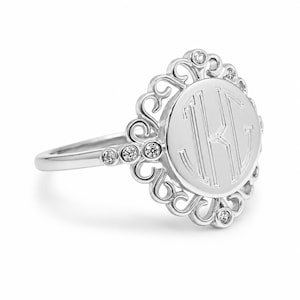 May include: A silver-toned ring featuring a circular face with an engraved monogram of the letters K and G. The circular face is surrounded by an ornate, openwork metal frame accented with small, clear, round-cut stones set along the border.