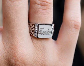 May include: A silver-toned signet ring featuring a rectangular face engraved with the name Heather in script font. The ring band displays a decorative cut-out pattern on the sides and is accented with small clear stones along the edges of the face.