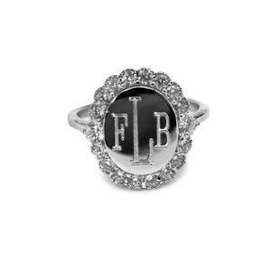 May include: A silver ring with a round, black, engraved center featuring the initials "FLB" surrounded by a halo of clear cubic zirconia stones.