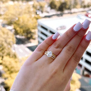 May include: A set of three gold rings. The engagement ring features a pear-shaped diamond. The other two rings are gold bands, one with small diamonds and the other a plain band. The rings are on a hand with light purple nails.