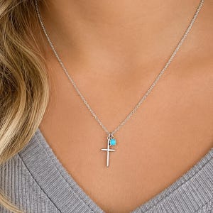 May include: A silver-colored necklace featuring a cross pendant and a small, round turquoise-colored bead. The cross is a simple, vertical design. The necklace is worn against a gray top.