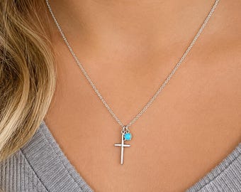 Könnte beinhalten: Eine silberfarbene Halskette mit einem Kreuzanhänger und einer kleinen, türkisfarbenen Perle. Das Kreuz hat ein einfaches, vertikales Design. Die Halskette wird über einem grauen Oberteil getragen.