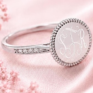 May include: A silver ring with a round, engraved face featuring the initials SFR. The ring has a rope-like border around the face and small clear stones set into the band. The ring is set against a soft pink background.