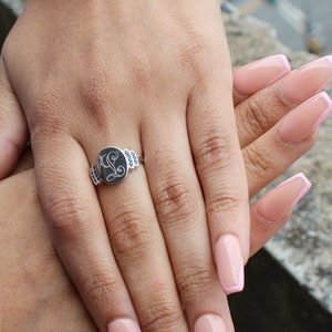 May include: A silver ring with a blue stone and a letter 'L' engraved on the face of the ring. The ring is on a person's finger with pink and white nail polish.