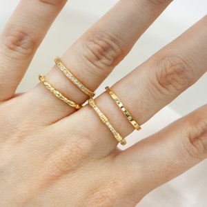 Puede incluir: Tres anillos de oro en una mano. Los anillos son de diferentes estilos, uno es una banda simple, otro tiene un diseño texturizado y el otro tiene pequeñas piedras transparentes.