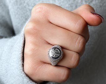 May include: A silver-toned signet ring featuring a circular face with an engraved monogram of the letters M, C, and G. The ring band and the area surrounding the circular face are decorated with a textured, granular pattern across the surface.