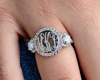 May include: A silver-toned ring featuring a central circular monogram plate with three intertwined script letters. The monogram is surrounded by a halo of small clear stones, flanked on each side by a round white opal stone set in silver.
