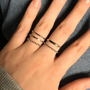 Puede incluir: Un conjunto de cuatro anillos de tono dorado usados en dos dedos. Dos anillos son bandas delgadas y lisas, un anillo presenta una serie de peque&ntilde;as piedras claras incrustadas y un anillo tiene una superficie texturizada de cuentas en el dedo medio.