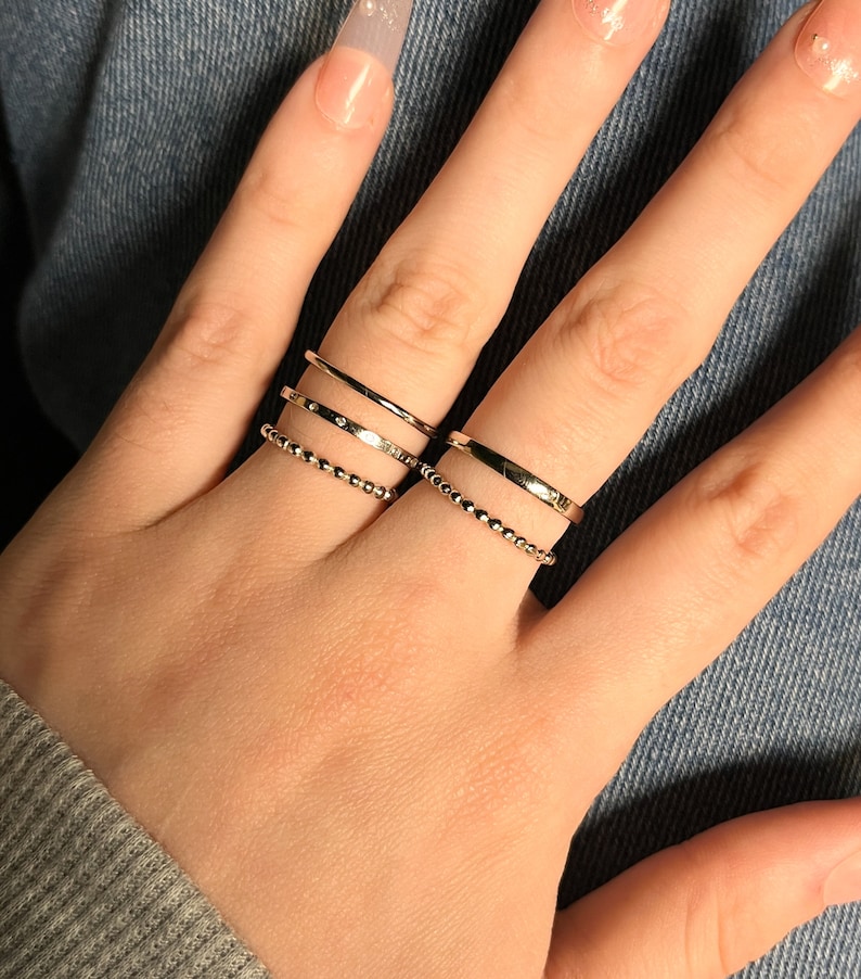 Puede incluir: Un conjunto de cuatro anillos de tono dorado usados en dos dedos. Dos anillos son bandas delgadas y lisas, un anillo presenta una serie de peque&ntilde;as piedras claras incrustadas y un anillo tiene una superficie texturizada de cuentas en el dedo medio.