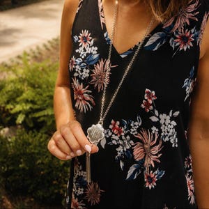 May include: Silver-toned necklace with a floral pendant and tassel detail. The pendant features an engraved monogram. The necklace is worn over a black floral print top. The background is blurred, with green foliage.