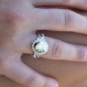May include: A silver ring with a round, clear stone setting. The stone has a monogram engraved on it. The ring is on a person's finger.