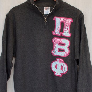 May include: A dark gray sweatshirt with a quarter zip closure. The sweatshirt has a pink and blue Greek letter design on the right side.