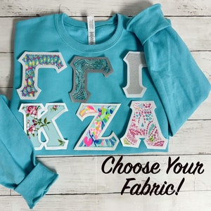 May include: A turquoise sweatshirt with a variety of fabric letters sewn on the front. The letters spell out "Gamma Zeta Alpha". The text "Choose Your Fabric!" is printed below the letters.