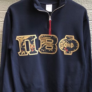 Op de afbeelding: Een donkerblauw sweatshirt met een geborduurd Grieks letterontwerp in goud. De letters zijn "&Pi;&Beta;&Phi;" en zijn omgeven door een bloemenpatroon.