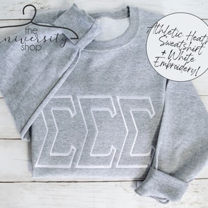 May include: Gray sweatshirt with white embroidered letters "&Sigma;&Sigma;&Sigma;" on the front. The sweatshirt is on a white wooden surface. The text "Athletic Heather Sweatshirt + White Embroidery!" is visible in the top right corner of the image.