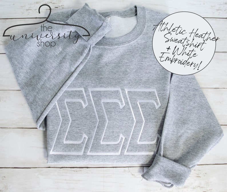 May include: Gray sweatshirt with white embroidered letters "&Sigma;&Sigma;&Sigma;" on the front. The sweatshirt is on a white wooden surface. The text "Athletic Heather Sweatshirt + White Embroidery!" is visible in the top right corner of the image.