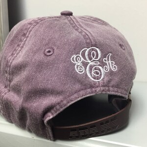 Op de afbeelding: Een paarse, verweerde, katoenen baseballpet met een wit geborduurd monogram. De pet heeft een bruin verstelbaar bandje.