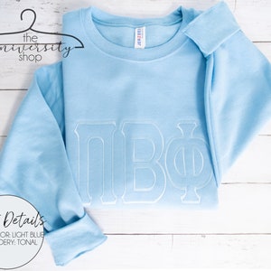 May include: Light blue sweatshirt with white embroidered Greek letters "&Pi;&Beta;&Phi;" on the front. "Shirt Details" is written in white text on a circle in the bottom left corner. "Shirt Color: Light Blue" and "Embroidery: Tonal" are written below the circle.
