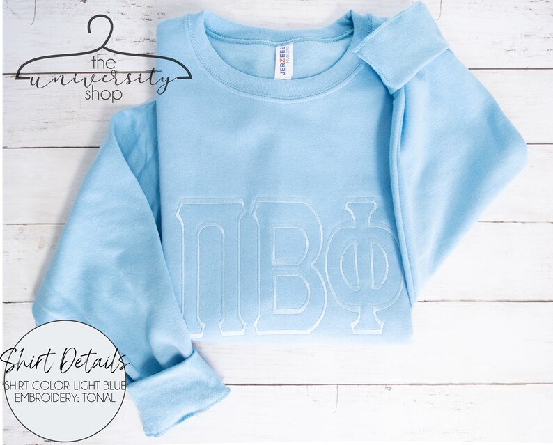 May include: Light blue sweatshirt with white embroidered Greek letters "&Pi;&Beta;&Phi;" on the front. "Shirt Details" is written in white text on a circle in the bottom left corner. "Shirt Color: Light Blue" and "Embroidery: Tonal" are written below the circle.