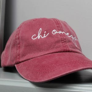 Op de afbeelding: Een bordeauxrode baseballcap van katoen met witte geborduurde tekst die "chi omega" luidt.