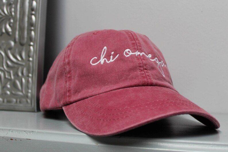 Op de afbeelding: Een bordeauxrode baseballcap van katoen met witte geborduurde tekst die "chi omega" luidt.