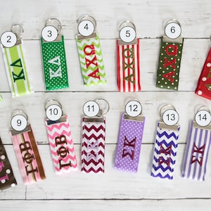 May include: Fifteen different keychains with various sorority and fraternity letters and patterns. The keychains are made of ribbon with different colors and patterns, including green, red, white, blue, purple, pink, and brown. The keychains are all attached to silver key rings.