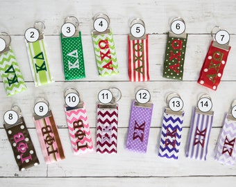 May include: Fifteen different keychains with various sorority and fraternity letters and patterns. The keychains are made of ribbon with different colors and patterns, including green, red, white, blue, purple, pink, and brown. The keychains are all attached to silver key rings.