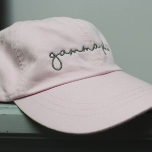 Op de afbeelding: Een roze baseballpet met het woord "gamma phi" in grijs geborduurd op de voorkant.