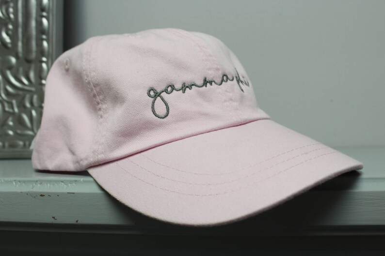 Op de afbeelding: Een roze baseballpet met het woord "gamma phi" in grijs geborduurd op de voorkant.
