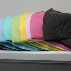 Op de afbeelding: Een stapel van 12 lege baseballpetten in verschillende pastelkleuren, waaronder roze, geel, groen, blauw en grijs. De petten zijn gerangschikt in een regenboogpatroon, met de donkerste kleur onderaan en de lichtste kleur bovenaan.