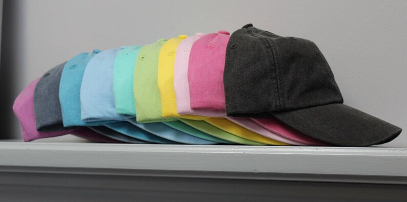 Op de afbeelding: Een stapel van 12 lege baseballpetten in verschillende pastelkleuren, waaronder roze, geel, groen, blauw en grijs. De petten zijn gerangschikt in een regenboogpatroon, met de donkerste kleur onderaan en de lichtste kleur bovenaan.