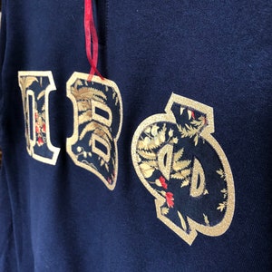 Op de afbeelding: Een marineblauw sweatshirt met een gouden geborduurd "IBO"-logo. De letters zijn omrand met goud en gevuld met een bloemenpatroon.