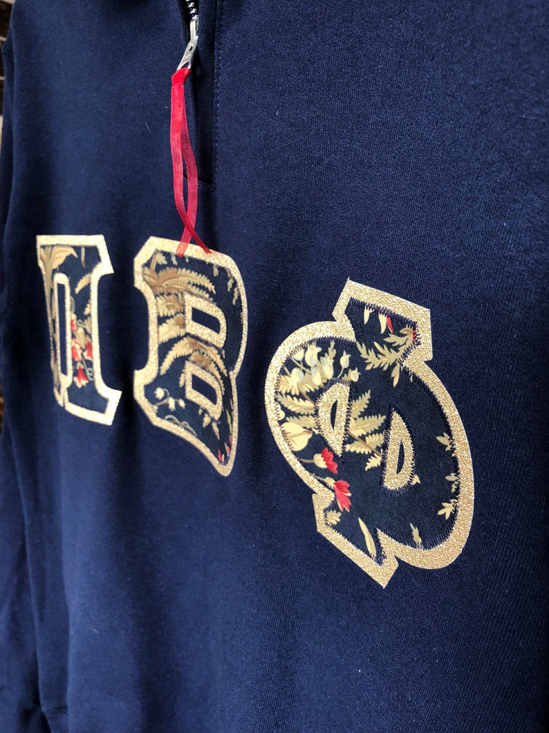 Op de afbeelding: Een marineblauw sweatshirt met een gouden geborduurd "IBO"-logo. De letters zijn omrand met goud en gevuld met een bloemenpatroon.