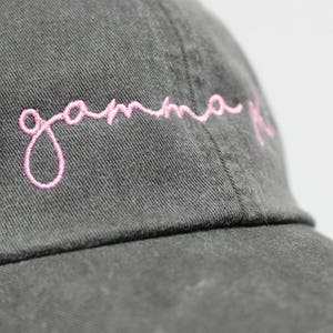 Op de afbeelding: Een donkergrijze baseballpet met een roze geborduurd cursief schrift dat "gamma phi" luidt.