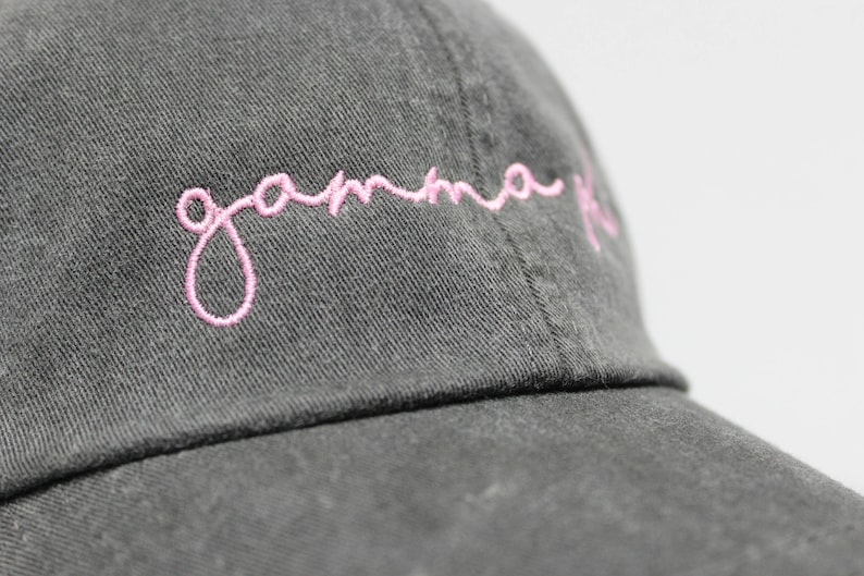 Op de afbeelding: Een donkergrijze baseballpet met een roze geborduurd cursief schrift dat "gamma phi" luidt.