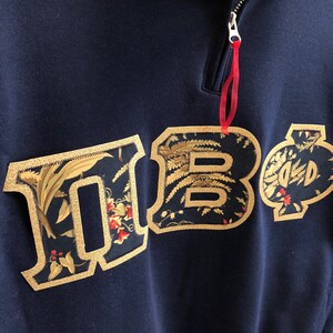 Op de afbeelding: Een marineblauw sweatshirt met een geborduurd bloemenpatroon in goud dat "HBO" spelt. Het sweatshirt heeft een ritssluiting bij de hals.