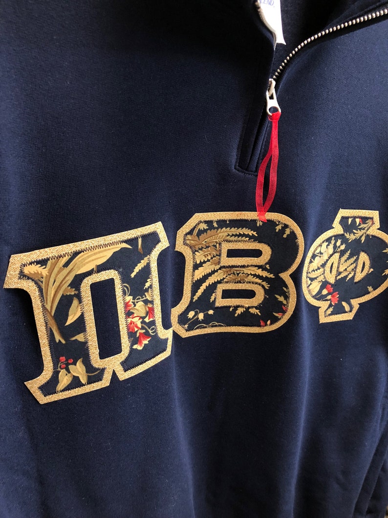 Op de afbeelding: Een marineblauw sweatshirt met een geborduurd bloemenpatroon in goud dat "HBO" spelt. Het sweatshirt heeft een ritssluiting bij de hals.