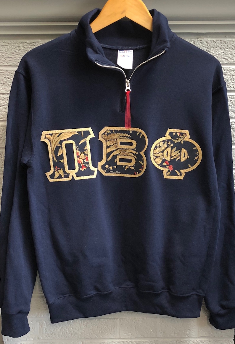 Op de afbeelding: Een donkerblauw sweatshirt met een geborduurd Grieks letterontwerp in goud. De letters zijn "&Pi;&Beta;&Phi;" en zijn omgeven door een bloemenpatroon.