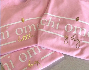 May include: Three pink t-shirts with white text and gold lettering. The text on the shirts reads "Chi Omega" with "little" and "big" written in cursive below the text.