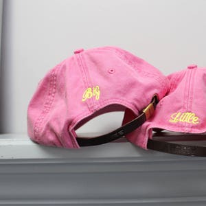 Op de afbeelding: Twee roze baseballpetten met verstelbare banden. De grotere pet heeft "Big" in geel geborduurd op de voorkant. De kleinere pet heeft "Litte" in geel geborduurd op de voorkant.
