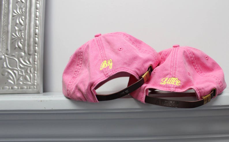 Op de afbeelding: Twee roze baseballpetten met verstelbare banden. De grotere pet heeft "Big" in geel geborduurd op de voorkant. De kleinere pet heeft "Litte" in geel geborduurd op de voorkant.